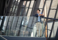 A businessman stands on a balcony of a corporate office, contemplating his professional reputation.