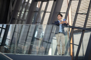 A businessman stands on a balcony of a corporate office, contemplating his professional reputation.
